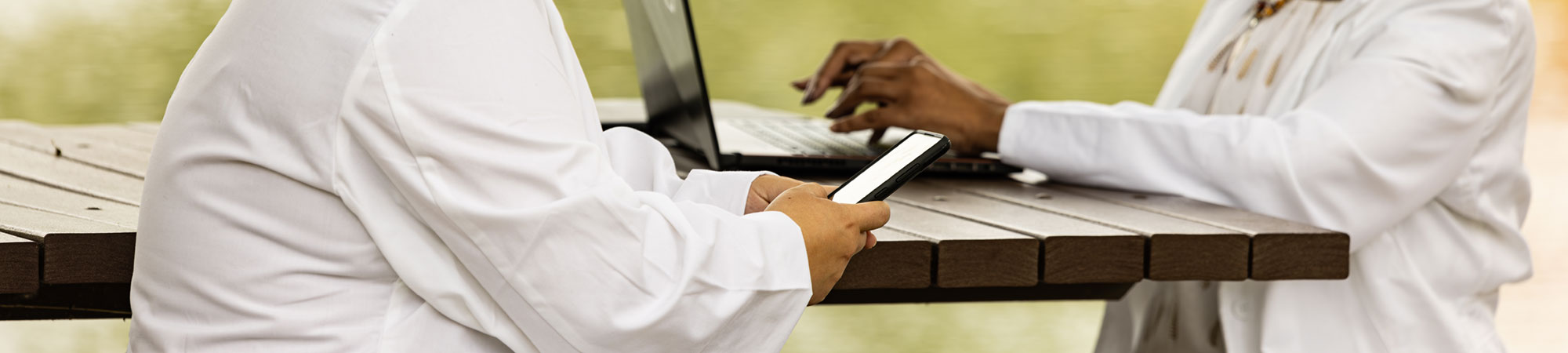 two students sitting at Lake Janse picnic tables on a phone and laptop